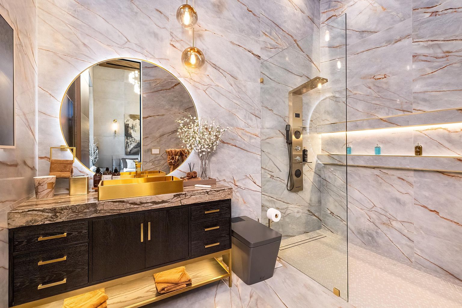 This luxurious bathroom features striking stone-clad walls with dramatic veining and a sleek, dark wood vanity topped with a metallic gold sink. A large, circular backlit mirror serves as a focal point, complemented by modern pendant lighting and a spacious walk-in shower enclosed by glass panels. The space exudes a high-end, contemporary aesthetic with gold accents and integrated shelf lighting that enhances the sophisticated atmosphere.