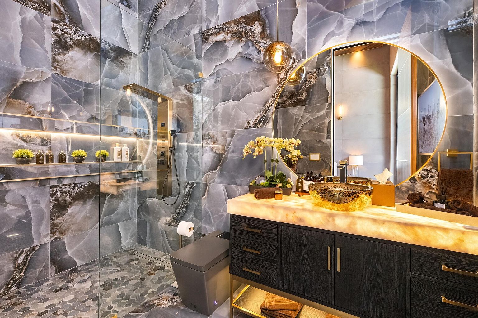 This luxurious bathroom features dramatic, large-format blue-toned stone wall tiles with striking veining that create a sophisticated, high-end aesthetic. The space includes a floating vanity with a glowing, backlit stone countertop, a unique gold-toned vessel sink, and a walk-in shower enclosed by glass panels. A large circular mirror with a gold frame reflects the space, while warm accent lighting under the vanity and within the shower niches adds a cozy, spa-like ambiance.