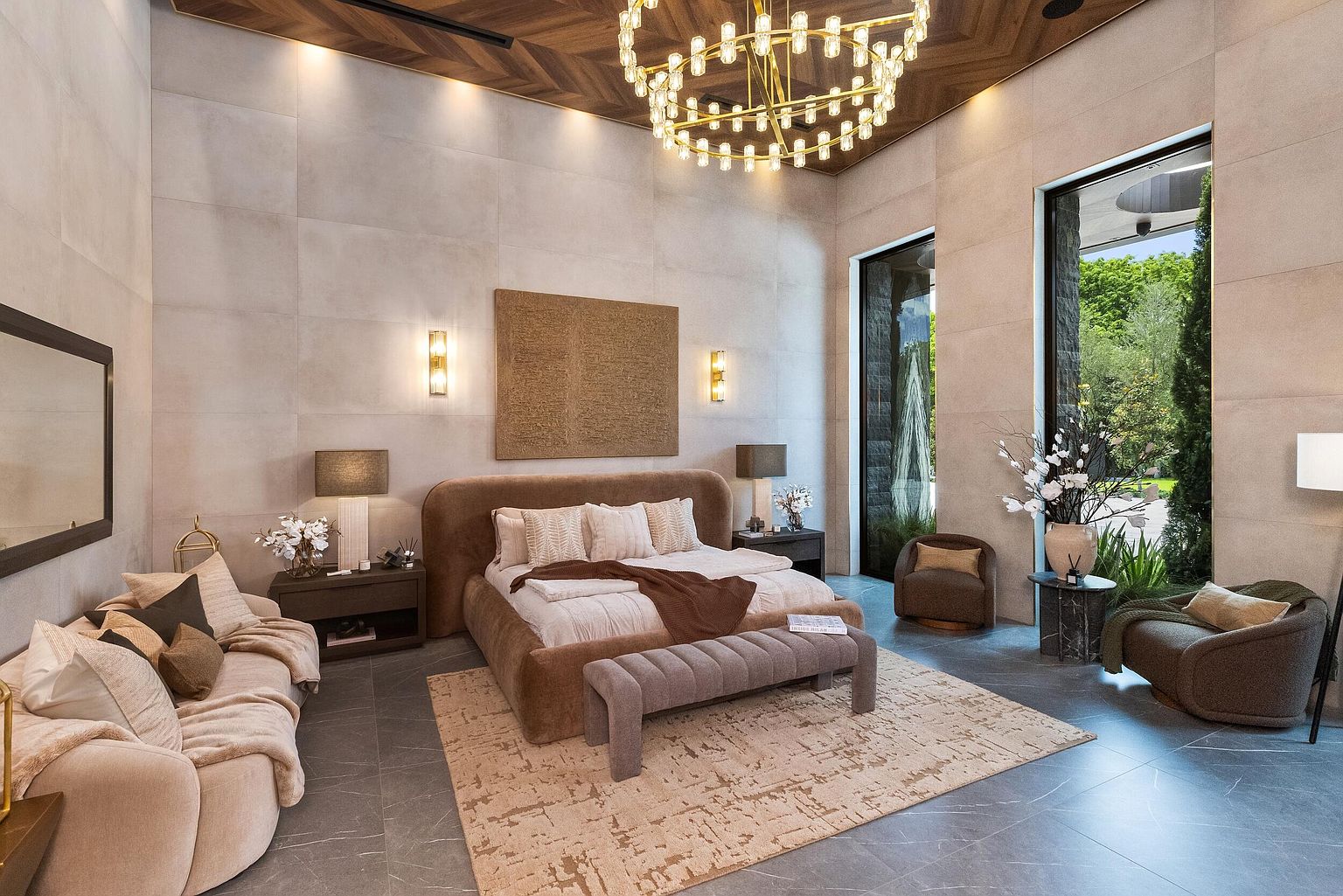 This expansive primary bedroom features a high, chevron-patterned wood ceiling and large windows that provide a view of the lush greenery outside. The room is anchored by a plush, rounded-edge bed with neutral bedding, complemented by a textured area rug and a modern, multi-tiered chandelier. A cozy seating area with a sofa and two armchairs creates a sophisticated, hotel-like atmosphere, while the neutral stone-tiled walls add a sense of refined elegance.