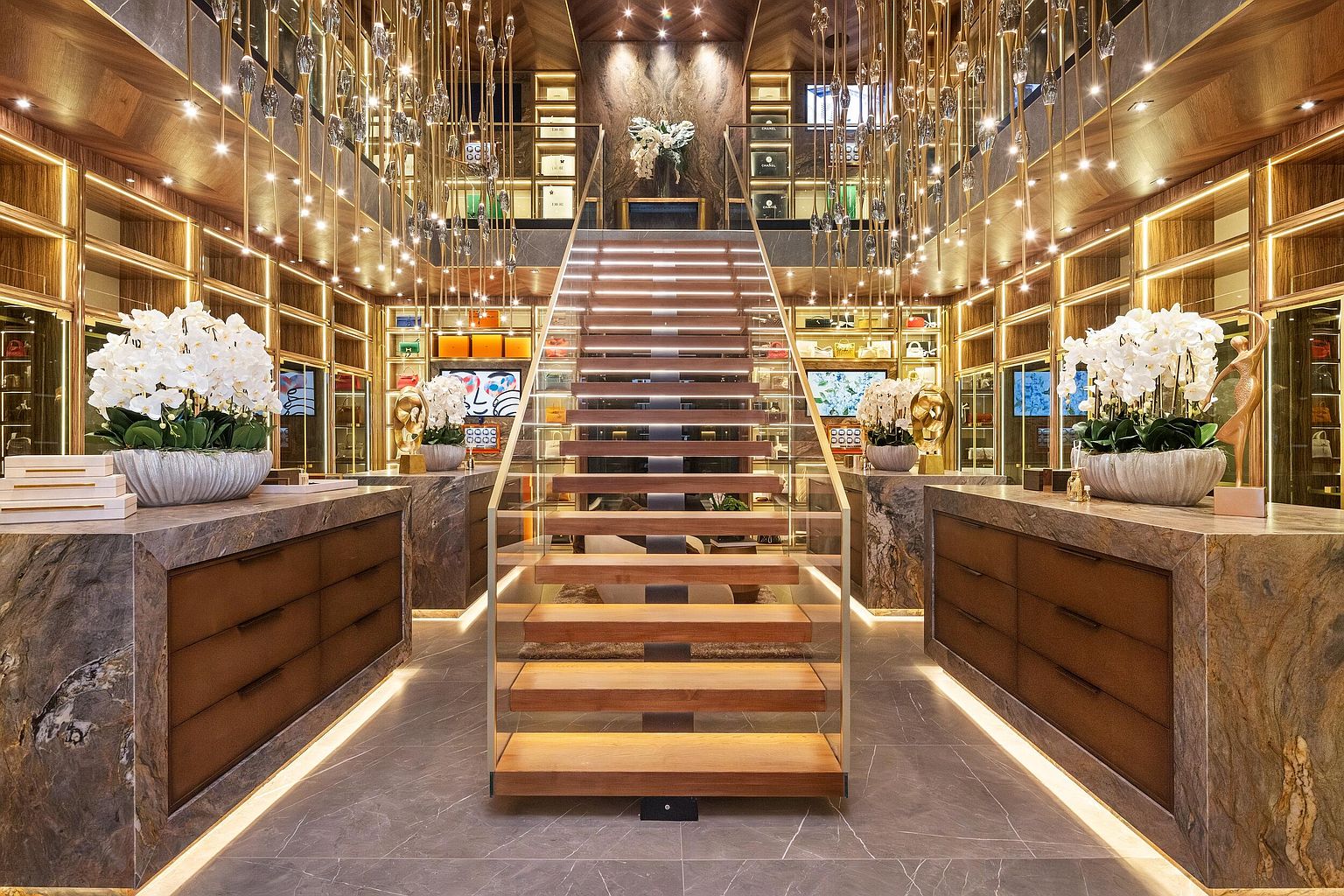 This luxurious, multi-level walk-in closet features a central floating staircase with wood treads and glass railings, serving as a dramatic focal point. The space is lined with extensive custom cabinetry, illuminated shelving for accessories, and large stone-topped islands on either side. Elegant hanging light fixtures descend from the ceiling, creating a sophisticated, boutique-like atmosphere.