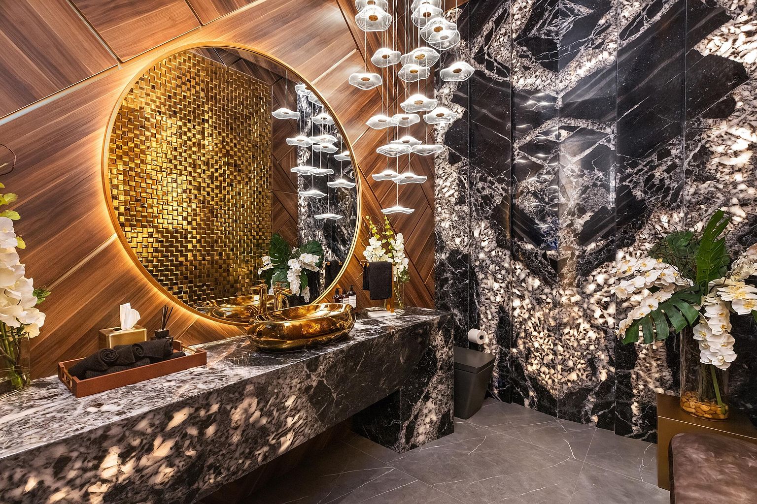 This luxurious powder room features a striking contrast between warm wood-paneled walls and dramatic, dark veined stone surfaces. A large, circular mirror with integrated backlighting hangs above a custom stone vanity, which is accented by a polished gold vessel sink and matching fixtures. A cascading, multi-tiered pendant light fixture adds a sophisticated, artistic touch to the space, while fresh floral arrangements enhance the opulent atmosphere.