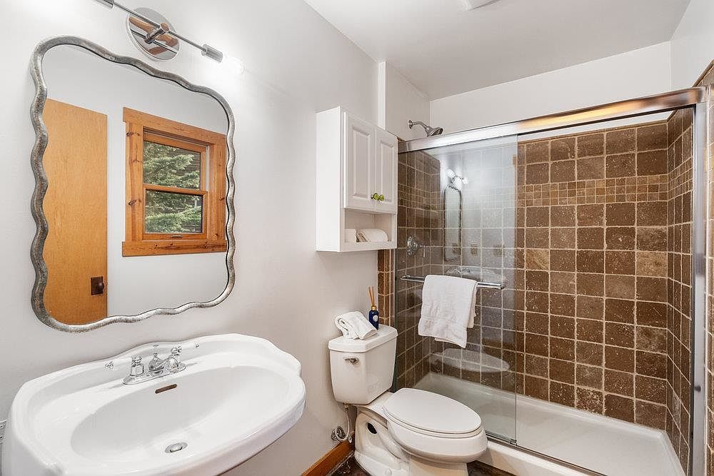 This is a bathroom featuring a white sink with a decorative mirror above it. A toilet is positioned next to a shower with brown tiled walls and a glass door. A white cabinet is mounted on the wall, providing storage space.