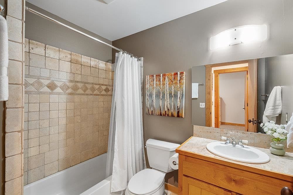 This is a bathroom featuring a bathtub with tiled walls and a shower curtain, a toilet, and a vanity with a sink. The walls are painted in a neutral tone, and there is artwork on the wall. The bathroom appears clean and well-maintained, with a functional layout.