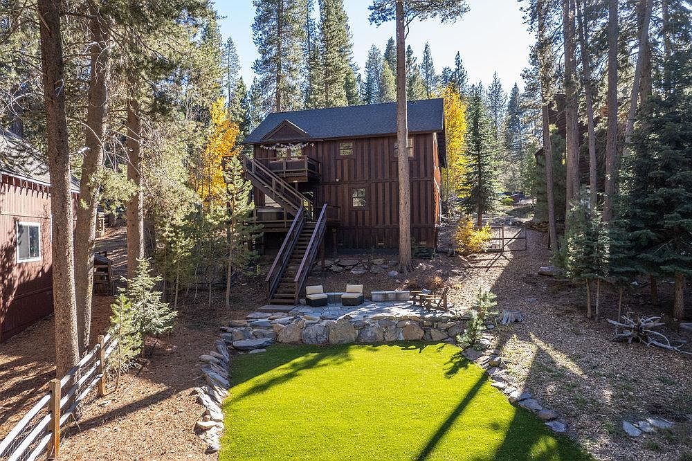 This image showcases a beautifully landscaped yard featuring a vibrant green lawn bordered by natural stone retaining walls. A wooden cabin-style house with exterior staircases is nestled among tall pine trees, creating a serene and private setting. The yard also includes a seating area with chairs and a small table, perfect for outdoor relaxation.