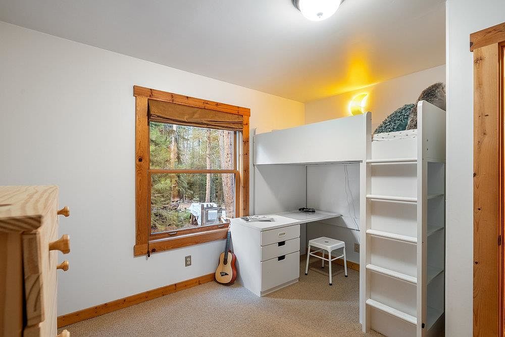 This is a cozy bedroom featuring a loft bed with a built-in desk underneath. The room has a warm and inviting feel, with natural light streaming in through a window framed by wood trim. A guitar leans against the wall, adding a touch of personality to the space.