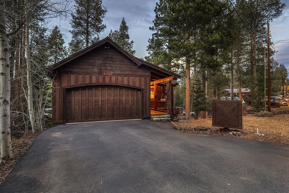 This image showcases the front exterior of a charming, rustic home. The house features a dark wood facade, a curved garage door, and a gabled roof. Tall trees surround the property, creating a sense of privacy and seclusion. The driveway is paved and leads up to the garage.