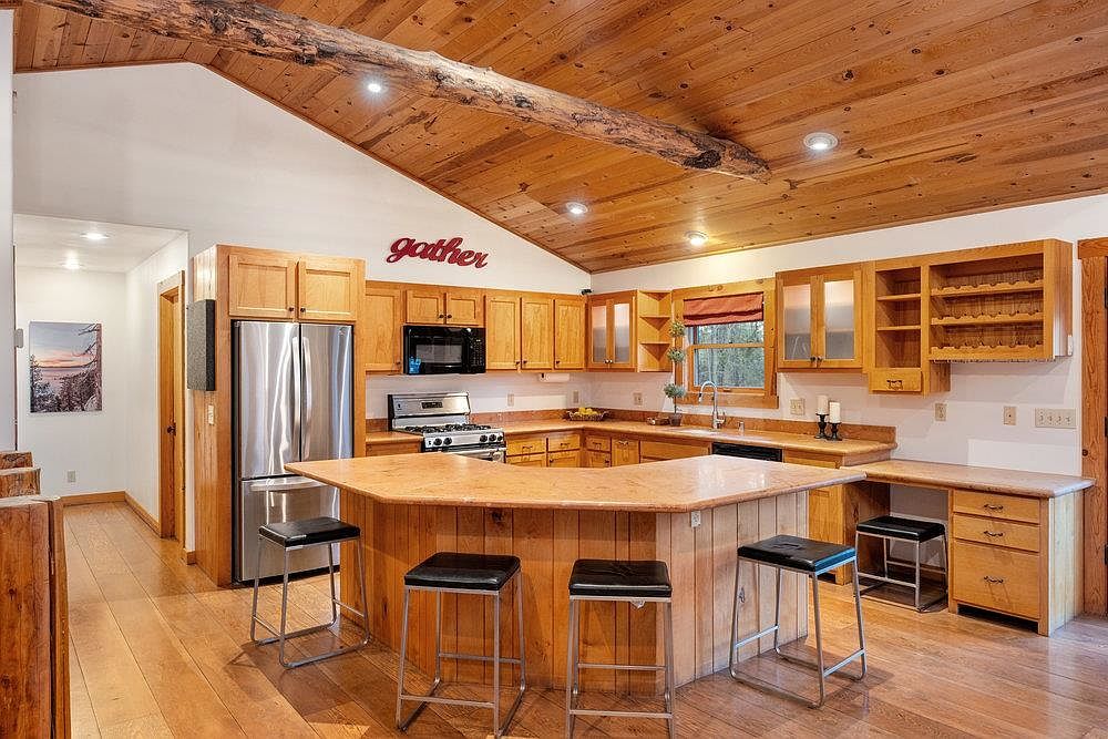 This is a warm and inviting kitchen with wood cabinetry, countertops, and flooring, creating a rustic and cozy atmosphere. A central island with bar stools provides seating, while stainless steel appliances offer modern convenience. The ceiling features wooden beams, adding to the room's character.