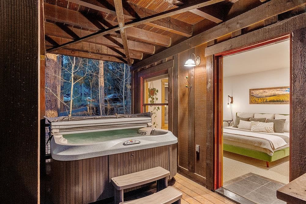 This image showcases a cozy outdoor patio area featuring a hot tub with wooden steps for access. The space is partially covered by a wooden structure, offering a sense of privacy and shelter. An open doorway leads into a bedroom, creating an indoor-outdoor flow.