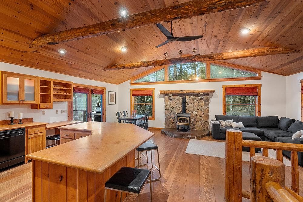 This interior shot showcases a cozy living room with a rustic aesthetic. The room features a stone fireplace, a dark gray sectional sofa, and wooden floors. The ceiling is clad in wood paneling with exposed beams, and natural light streams in through the windows, creating a warm and inviting atmosphere.