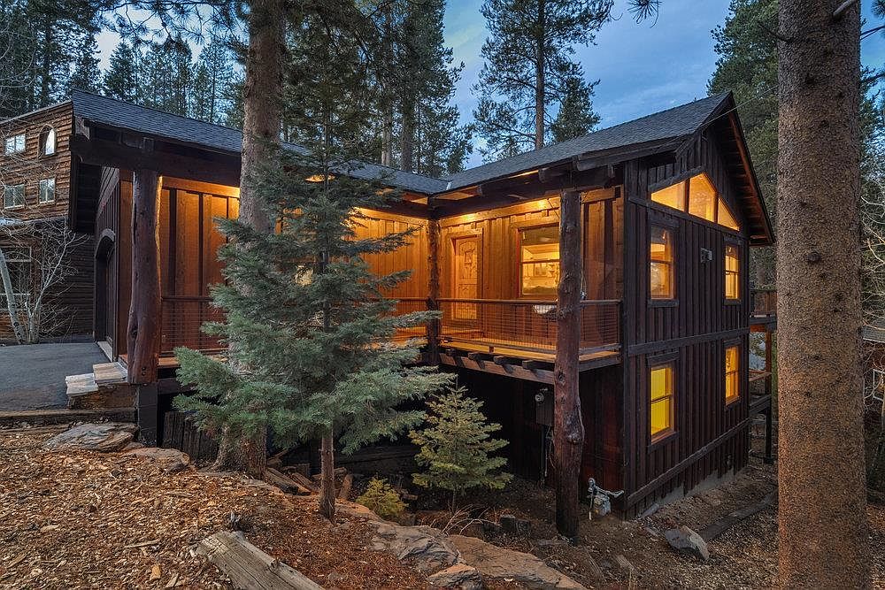 This is a side view of a rustic, multi-level cabin nestled among tall trees. The exterior features dark wood siding, a deck with railing, and several windows that glow with warm interior light. The surrounding landscape includes natural ground cover and mature trees, enhancing the cabin's secluded and cozy atmosphere.