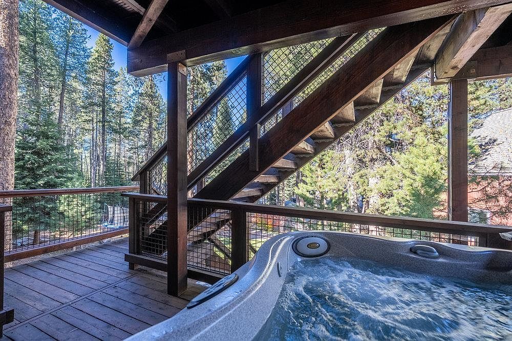 This image showcases a wooden deck featuring a built-in hot tub with bubbling water. The deck is surrounded by a wooden railing with metal mesh, offering a view of the surrounding forest. A wooden staircase leads to another level, adding architectural interest and suggesting multiple outdoor living spaces.