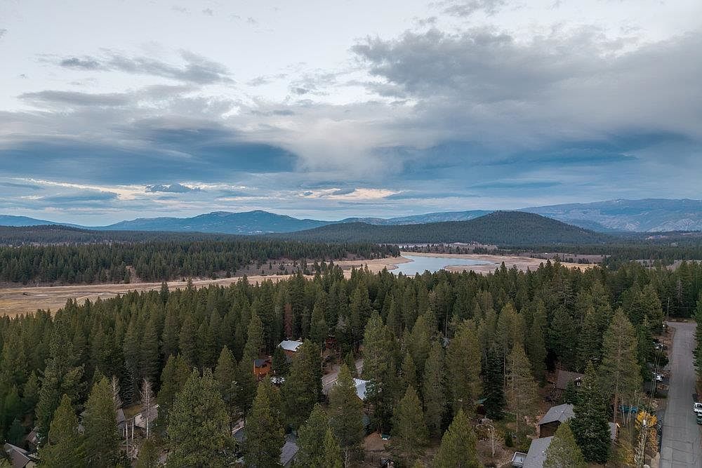This aerial view showcases a vast landscape featuring a dense forest, a serene lake, and distant mountains under a cloudy sky. The presence of houses nestled among the trees suggests a peaceful residential area, highlighting the property's proximity to nature and potential for a tranquil lifestyle. A road winds through the trees, indicating accessibility.