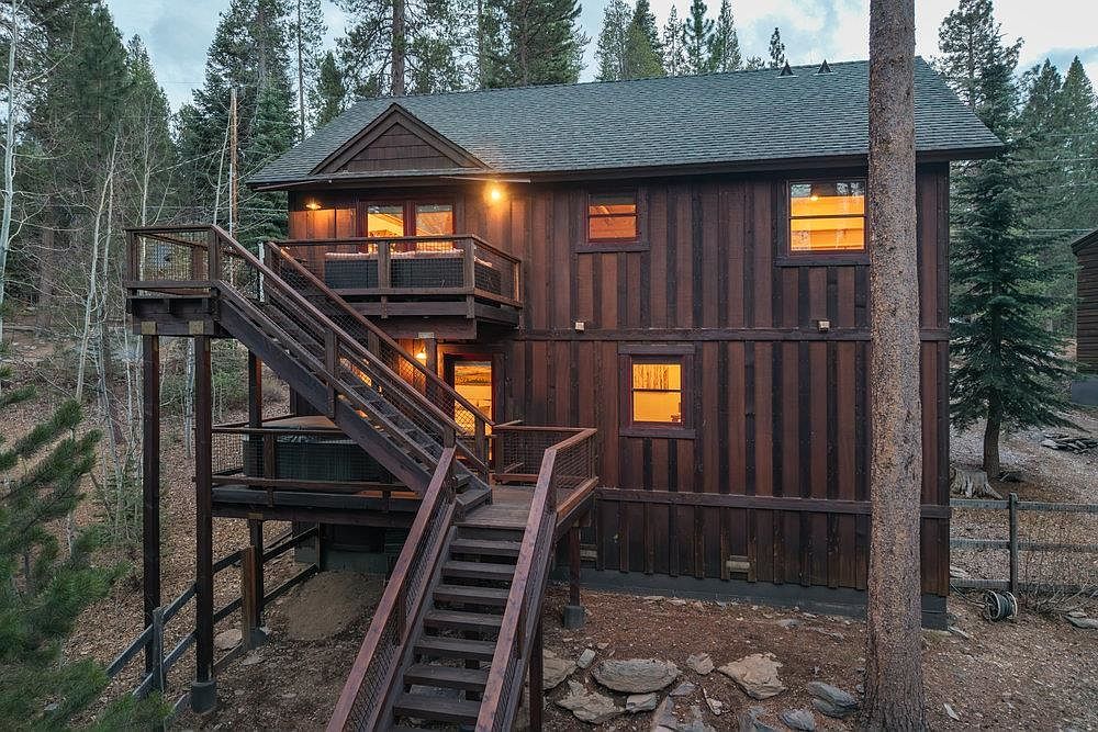 This is a front exterior view of a multi-story cabin-style home with dark wood siding and a green roof. The house features multiple decks accessible by wooden staircases, creating an inviting outdoor living space. Windows are illuminated, suggesting a warm and cozy interior, while the surrounding trees provide a sense of privacy and seclusion.