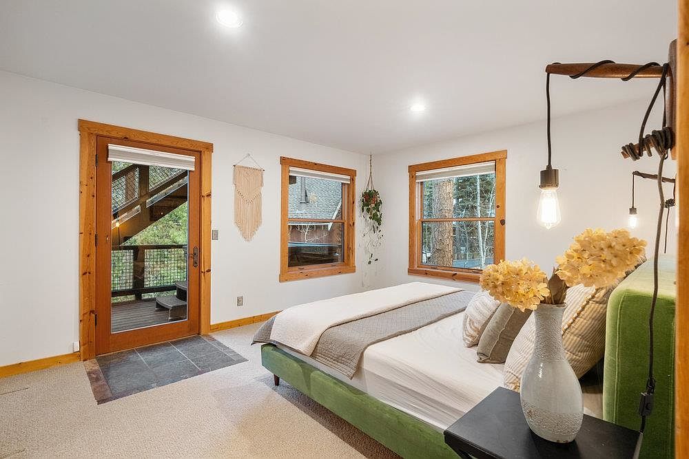 This is a cozy primary bedroom featuring a green bed frame, neutral bedding, and wooden-framed windows that provide natural light. The room has a rustic-modern aesthetic, with a unique wooden lamp fixture and a macrame wall hanging adding to the charm. A door leads to an outdoor deck, enhancing the indoor-outdoor flow.