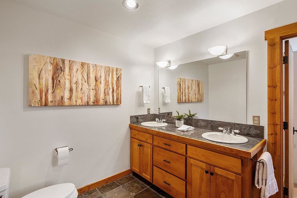 This is a well-lit primary bathroom featuring a double vanity with a dark countertop and wooden cabinets. A large mirror reflects the space, and artwork hangs on the wall, adding a touch of nature. The overall impression is clean and functional with a rustic touch.