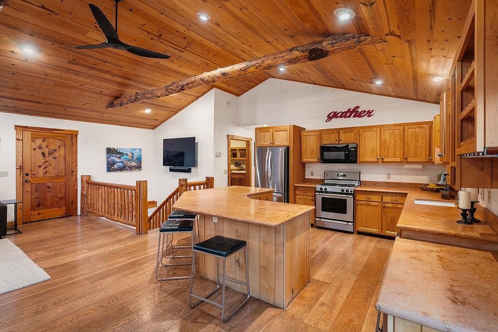 This interior shot showcases a warm and inviting kitchen with wood cabinetry, countertops, and flooring, complemented by a unique wooden beam on the ceiling. Stainless steel appliances and a kitchen island with bar seating add functionality, while the overall design exudes a rustic and cozy atmosphere. The room also features a staircase and a door leading to another room.