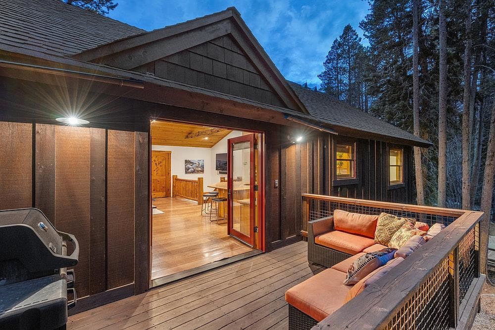 This image showcases a cozy outdoor deck area attached to a rustic cabin-style home. The deck features comfortable seating with orange cushions and decorative pillows, enclosed by a wooden railing with metal mesh. An open doorway leads into the home, revealing a glimpse of the interior with hardwood floors and wooden accents, creating an inviting indoor-outdoor living space.