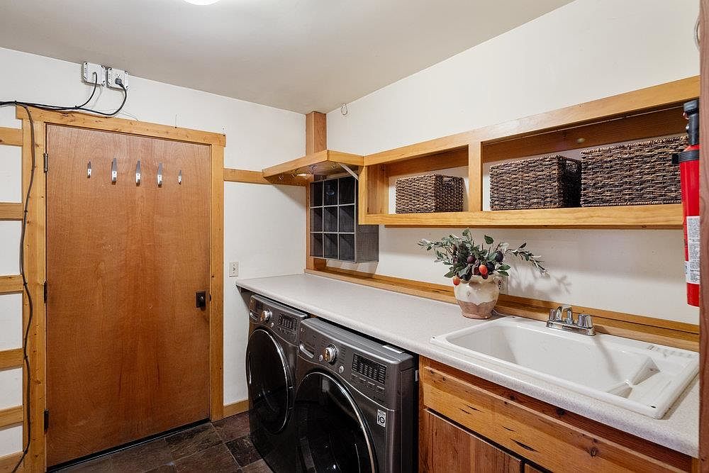 This is an interior shot of a laundry room featuring a front-loading washer and dryer set, a utility sink with wooden cabinetry, and open wooden shelving with decorative baskets. A wooden door with hooks is visible on the left, and the room has a rustic, functional aesthetic with a focus on practical storage solutions.