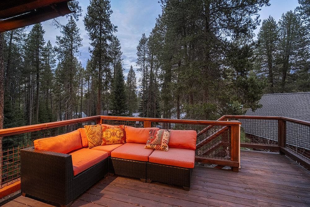 This image showcases a cozy outdoor living space on a wooden deck, featuring a sectional sofa with orange cushions and decorative pillows. The deck is surrounded by a wooden railing with wire mesh, offering views of the surrounding forest. The scene evokes a sense of relaxation and connection with nature, perfect for enjoying the outdoors.