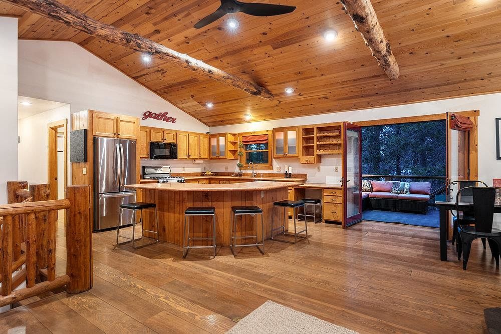 This is a warm and inviting kitchen with wood cabinetry, stainless steel appliances, and a large central island with bar seating. The wood-paneled ceiling with exposed beams adds a rustic touch, while the open doorway leads to an outdoor seating area, creating a seamless indoor-outdoor flow. The overall impression is cozy and well-suited for entertaining.