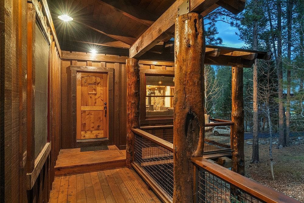 This image showcases a rustic-style patio or deck area, featuring wooden construction with log pillars and metal railing. The space is well-lit with overhead lighting, highlighting the natural wood grain and the surrounding trees. The overall impression is one of cozy, outdoor living with a touch of nature.