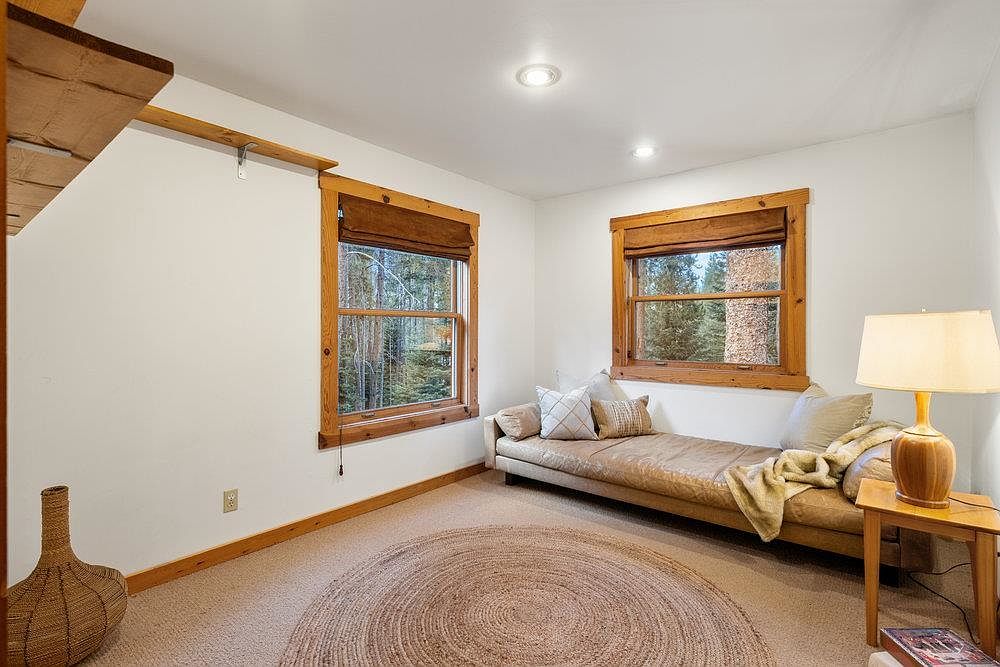 This is a cozy bedroom featuring two windows with wooden frames and brown shades, allowing natural light to filter in. A daybed with neutral-toned cushions and pillows sits against one wall, complemented by a wooden side table and a lamp. A circular jute rug adds texture to the carpeted floor, creating a warm and inviting atmosphere.