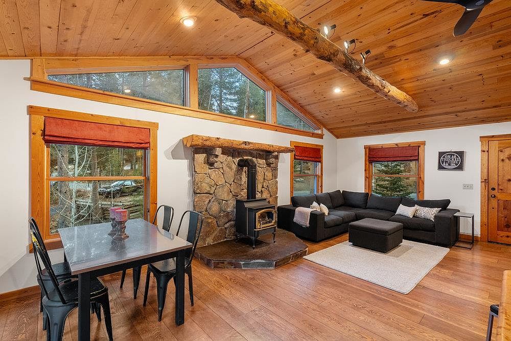 This inviting living room features a cozy stone fireplace with a wood-burning stove, complemented by warm wood ceilings and flooring. A comfortable sectional sofa and a neutral rug create a relaxing atmosphere. Natural light streams in through the windows, enhancing the room's appeal.