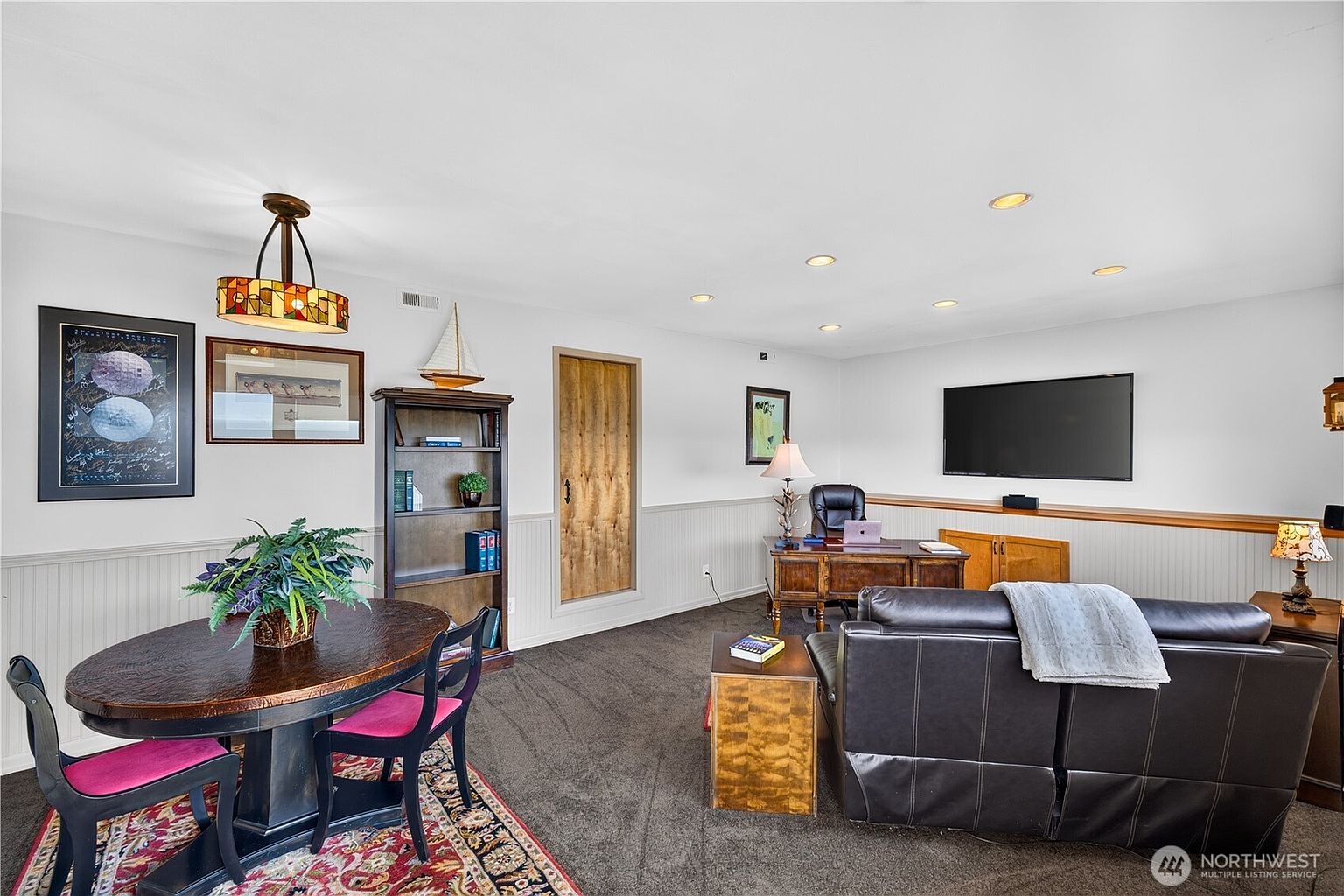 This multi-functional room serves as both a home office and a dining area, featuring dark carpeting and white wainscoting along the walls. The space includes a wooden desk with an office chair, a television mounted on the wall, and a round dining table with chairs set atop a patterned rug. A tall bookshelf and a decorative wooden door add character to the room, which is illuminated by recessed lighting and a stained-glass pendant fixture.