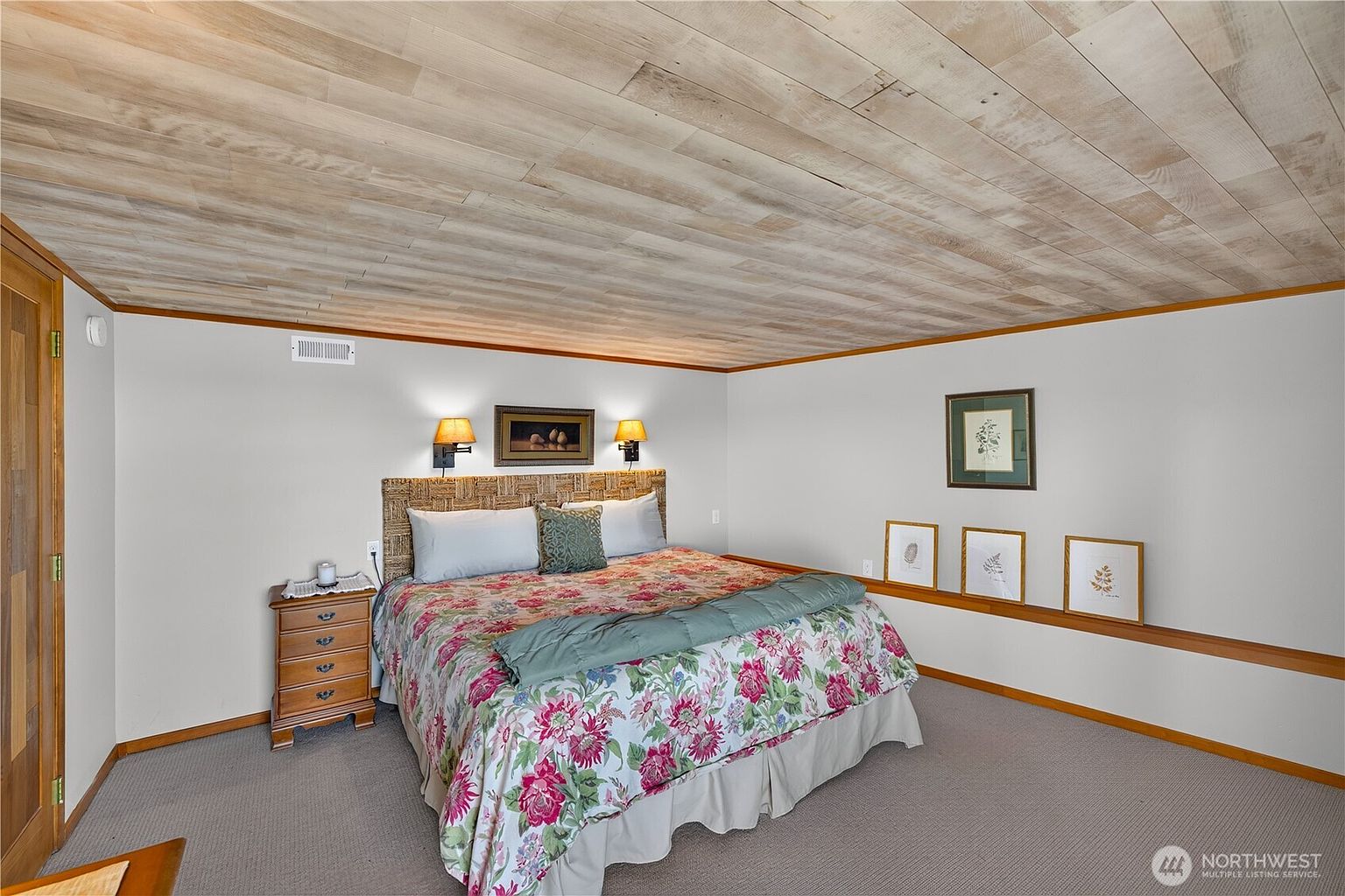 This bedroom features a large bed with a floral patterned comforter and a woven headboard, flanked by two wall-mounted lamps. The room is characterized by a light wood-paneled ceiling and neutral walls, with a series of framed botanical prints displayed along a low ledge. The space is carpeted and includes a wooden nightstand, creating a cozy and traditional atmosphere.