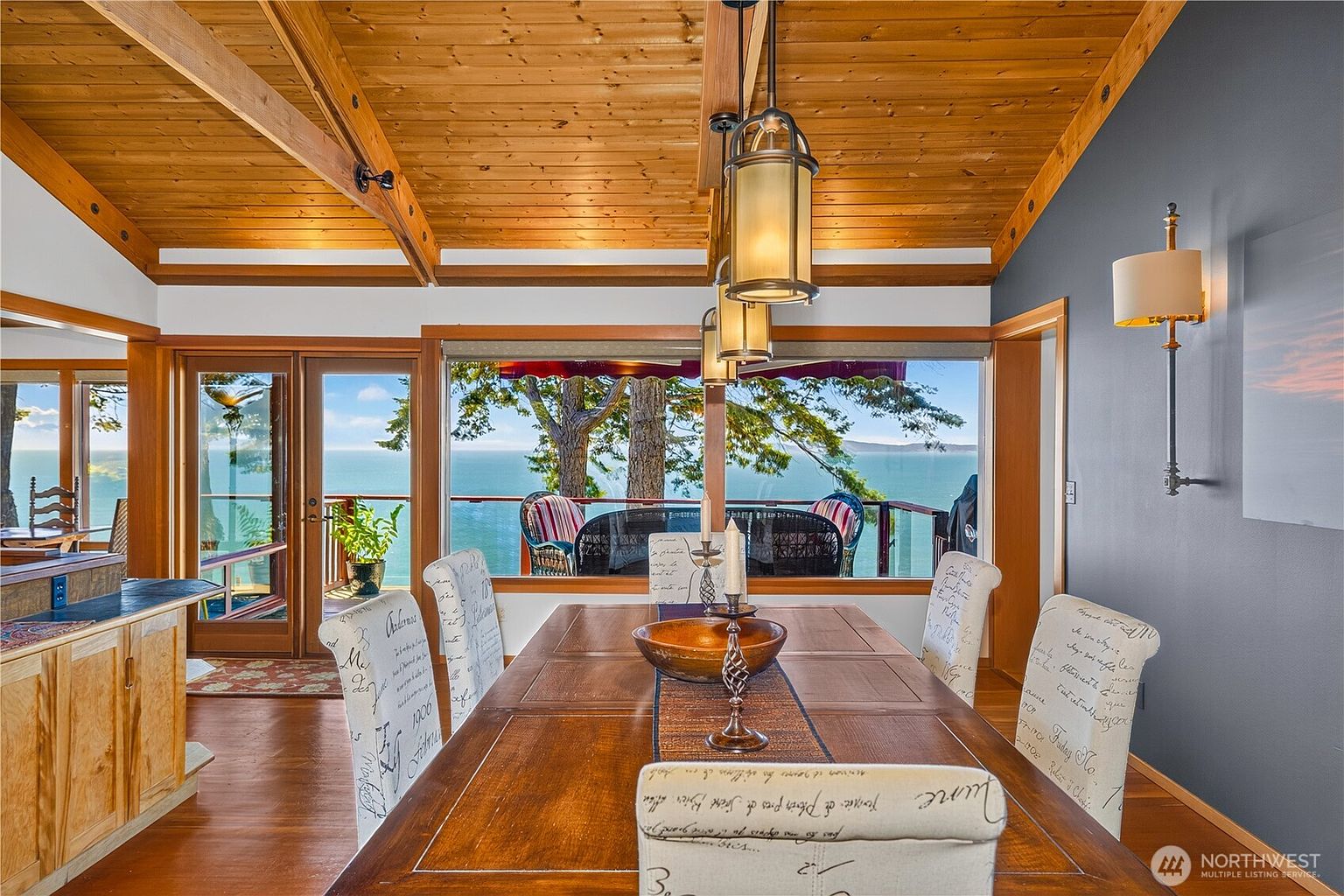 This dining room features a large wooden table surrounded by chairs with patterned fabric upholstery, set beneath a series of hanging pendant lights. The room is defined by a vaulted wood-paneled ceiling and a large window wall that provides an expansive, unobstructed view of the ocean and deck. A dark gray accent wall with a wall-mounted light fixture adds a modern touch to the warm, rustic atmosphere.