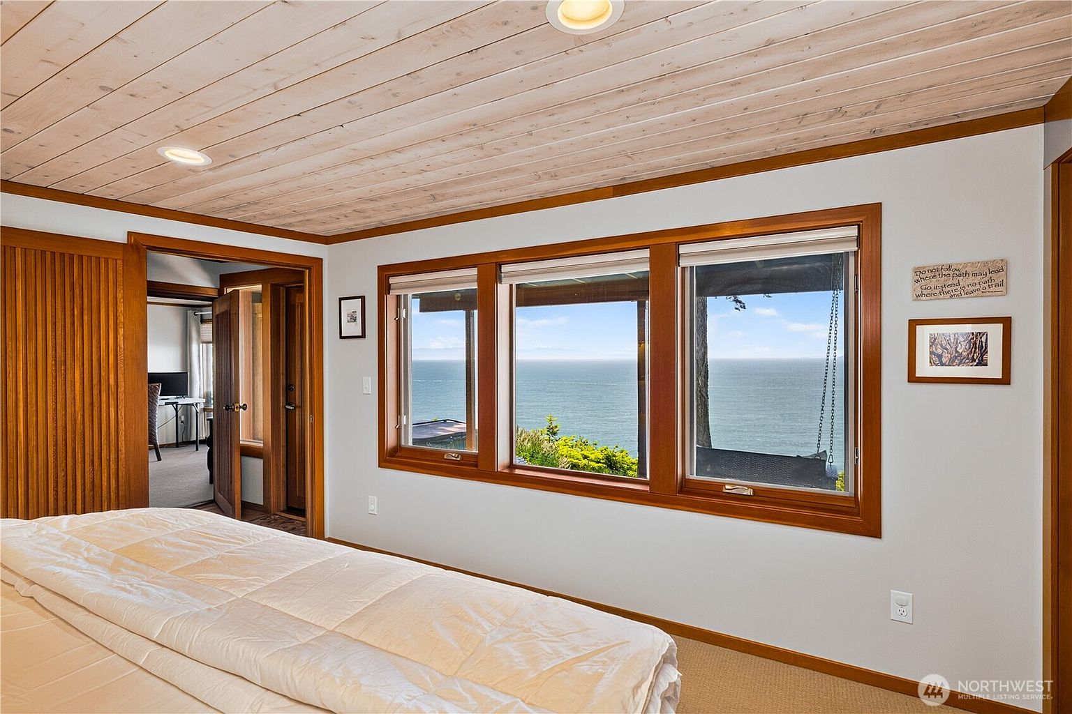 This bedroom features a light wood-paneled ceiling and a large set of windows that frame a panoramic view of the ocean. The room is neutrally decorated with a white bedspread and light-toned walls, accented by warm wood trim around the windows and doorways. A doorway leads into an adjacent room, providing a sense of depth and connectivity within the home.
