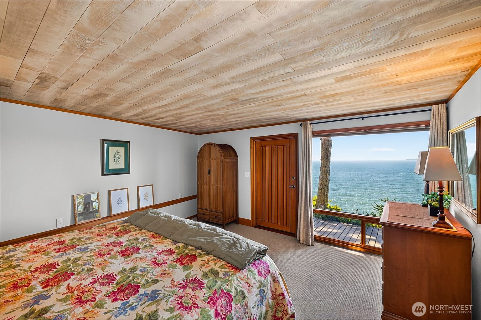 This bedroom features a distinctive light wood plank ceiling and a large window that frames a panoramic view of the ocean. The room is furnished with a bed covered in a vibrant floral quilt, a wooden wardrobe, and a dresser topped with a lamp. The space offers a serene, coastal atmosphere with direct access to an outdoor deck through a wooden door.
