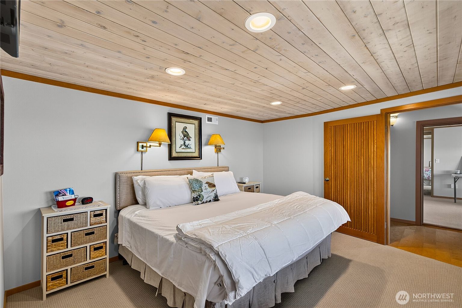 This primary bedroom features a light-toned wood plank ceiling with recessed lighting and neutral-colored walls. A large bed with a tufted headboard serves as the focal point, flanked by two wall-mounted lamps and a framed bird print. To the left, a small chest with woven baskets provides storage, while a wooden door leads to an adjacent room, creating a cozy and well-lit living space.