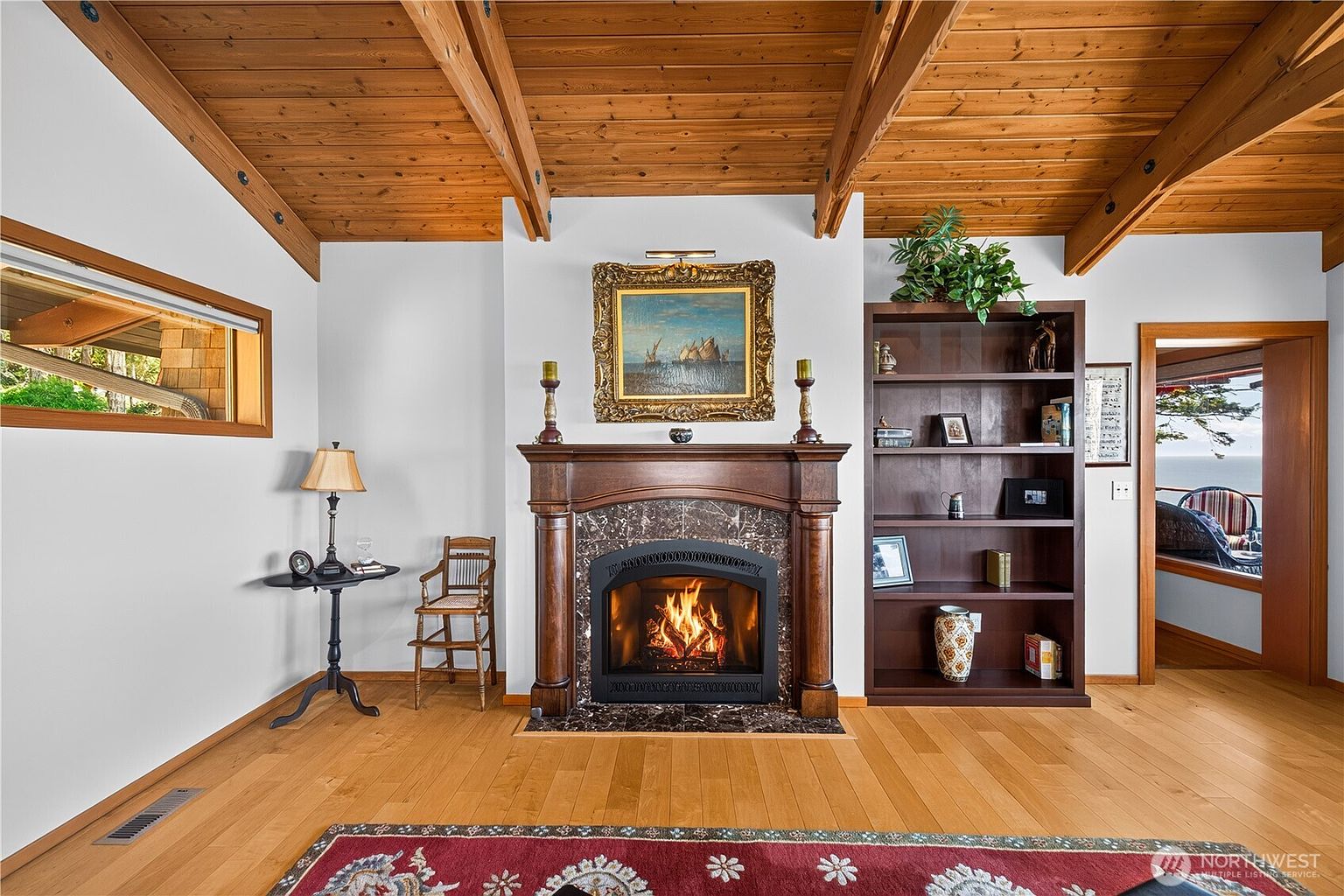 This living room features a prominent fireplace with a dark wood mantel and a stone surround, serving as the central focal point beneath a vaulted wood-paneled ceiling. To the right, a tall, dark wood bookshelf provides ample storage, while a small side table and chair are positioned to the left. The room is finished with light wood flooring and a large, patterned area rug, creating a warm and inviting atmosphere.