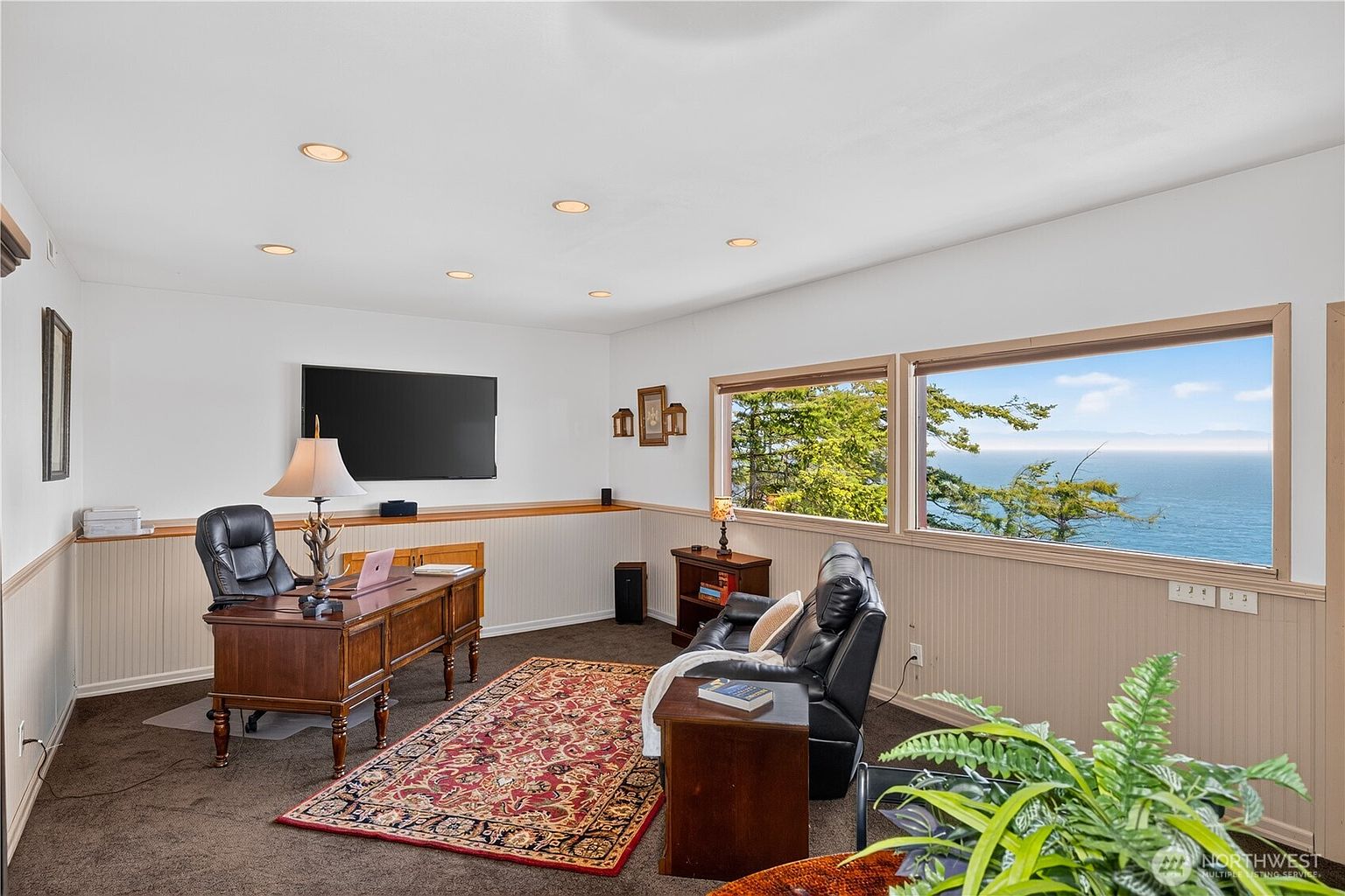 This home office features a traditional wooden desk, a black leather executive chair, and a matching black leather recliner positioned to enjoy the expansive ocean views through large windows. The room is finished with light-colored wainscoting, dark carpeting, and a decorative patterned area rug, creating a professional yet comfortable workspace. A wall-mounted television and recessed lighting complete the functional layout of this serene, view-oriented room.