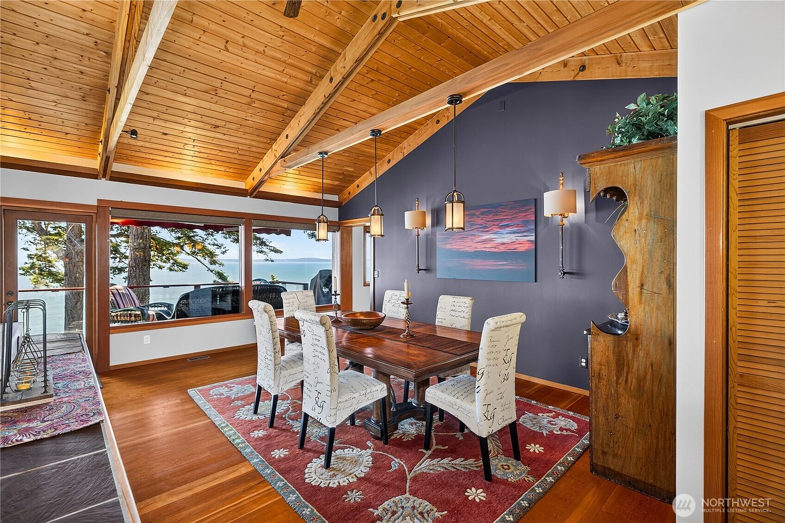 This dining room features a vaulted wood-paneled ceiling with exposed beams and a large window offering a scenic view of the water. The space is anchored by a dark wood dining table surrounded by chairs with patterned fabric, all set atop a vibrant red area rug. A dark accent wall serves as a backdrop for wall-mounted sconces and a piece of landscape art, while a tall wooden hutch adds a rustic touch to the room.