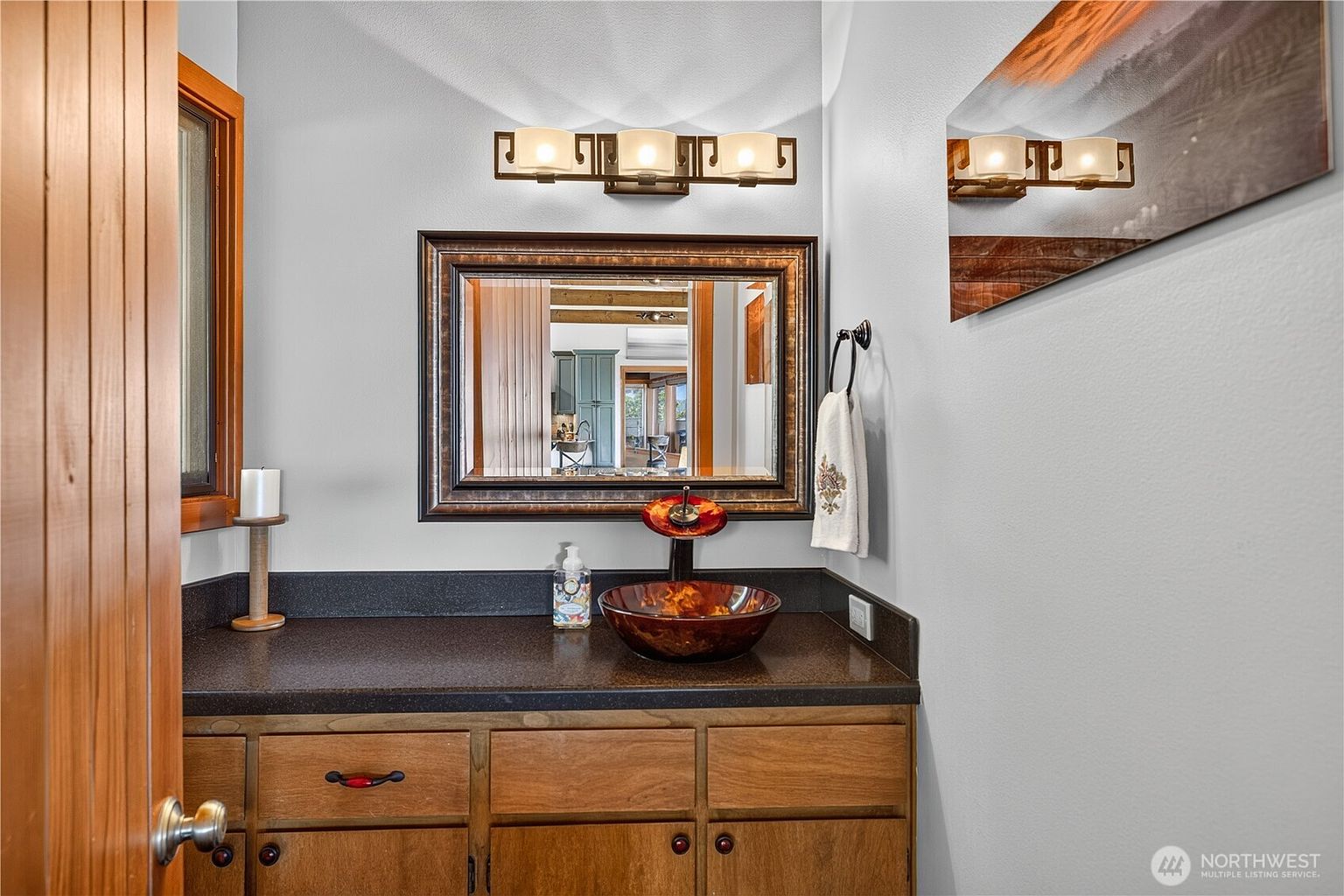 This bathroom features a rustic vanity with a dark countertop and a unique, patterned glass vessel sink. A framed mirror hangs above the sink, flanked by a three-light vanity fixture, while a hand towel hangs on a ring to the right. The space is finished with light-colored walls and wood-trimmed accents, creating a warm and cozy atmosphere.