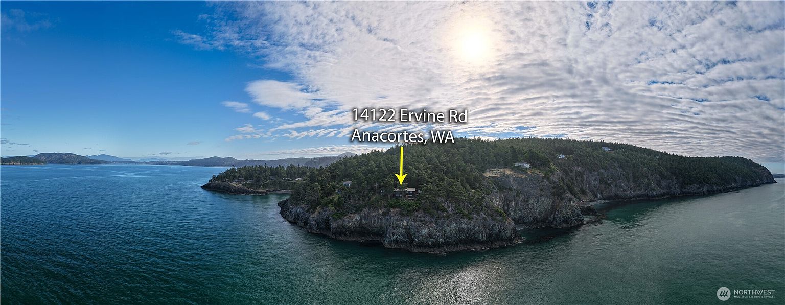 This wide-angle aerial shot captures a secluded coastal property situated on a rocky cliffside overlooking the water. The home is nestled within a dense forest of evergreen trees, emphasizing its private and scenic location. The perspective provides a panoramic view of the surrounding coastline, highlighting the dramatic topography and the expansive ocean horizon.