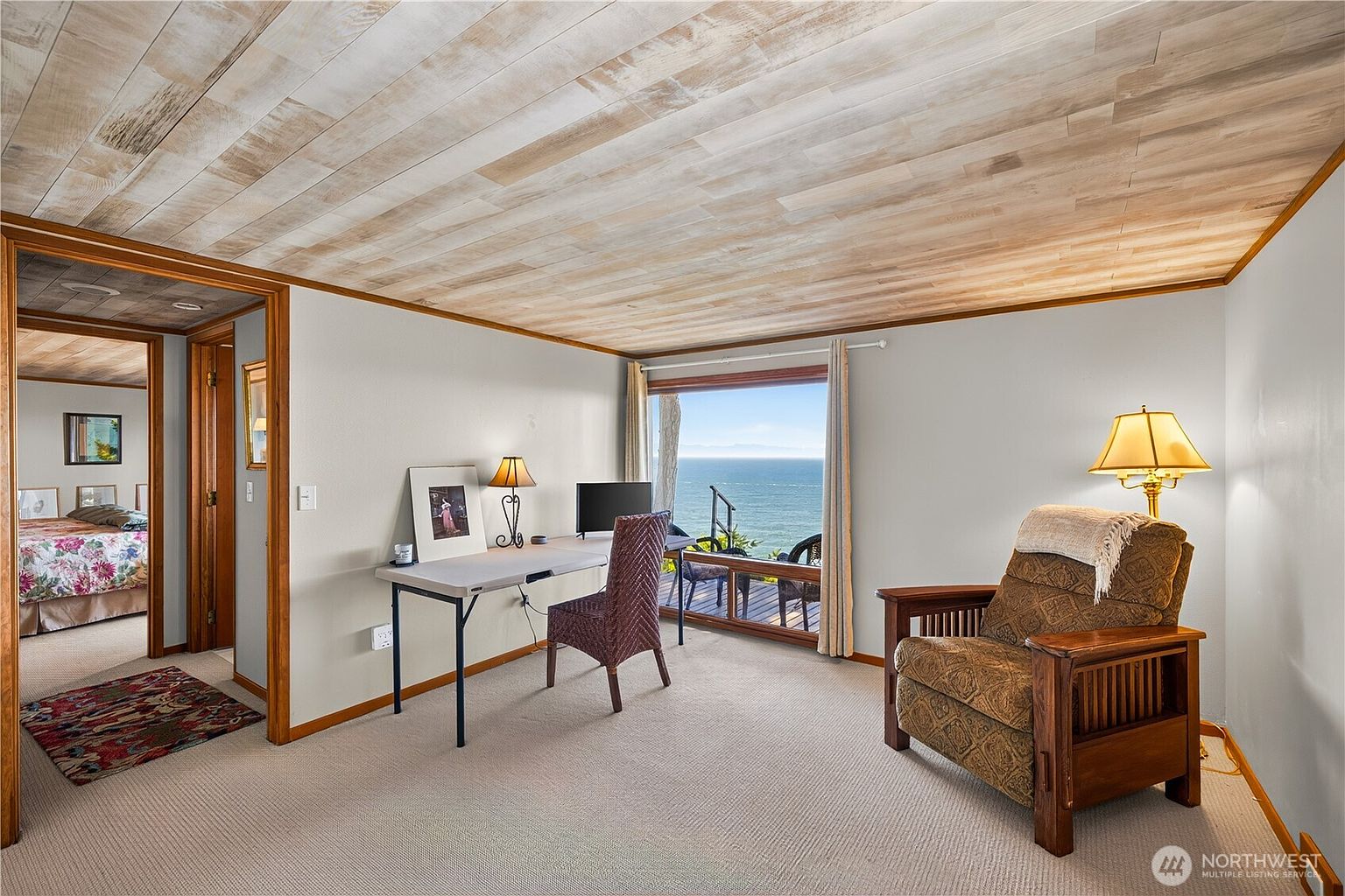 This bright office space features a light wood-paneled ceiling and neutral walls, creating a calm and focused atmosphere. A desk is positioned to take advantage of a large window that offers a stunning, unobstructed view of the ocean and a wooden deck. To the right, a comfortable upholstered armchair with a lamp provides a cozy reading nook, while a doorway leads into a bedroom with floral bedding.