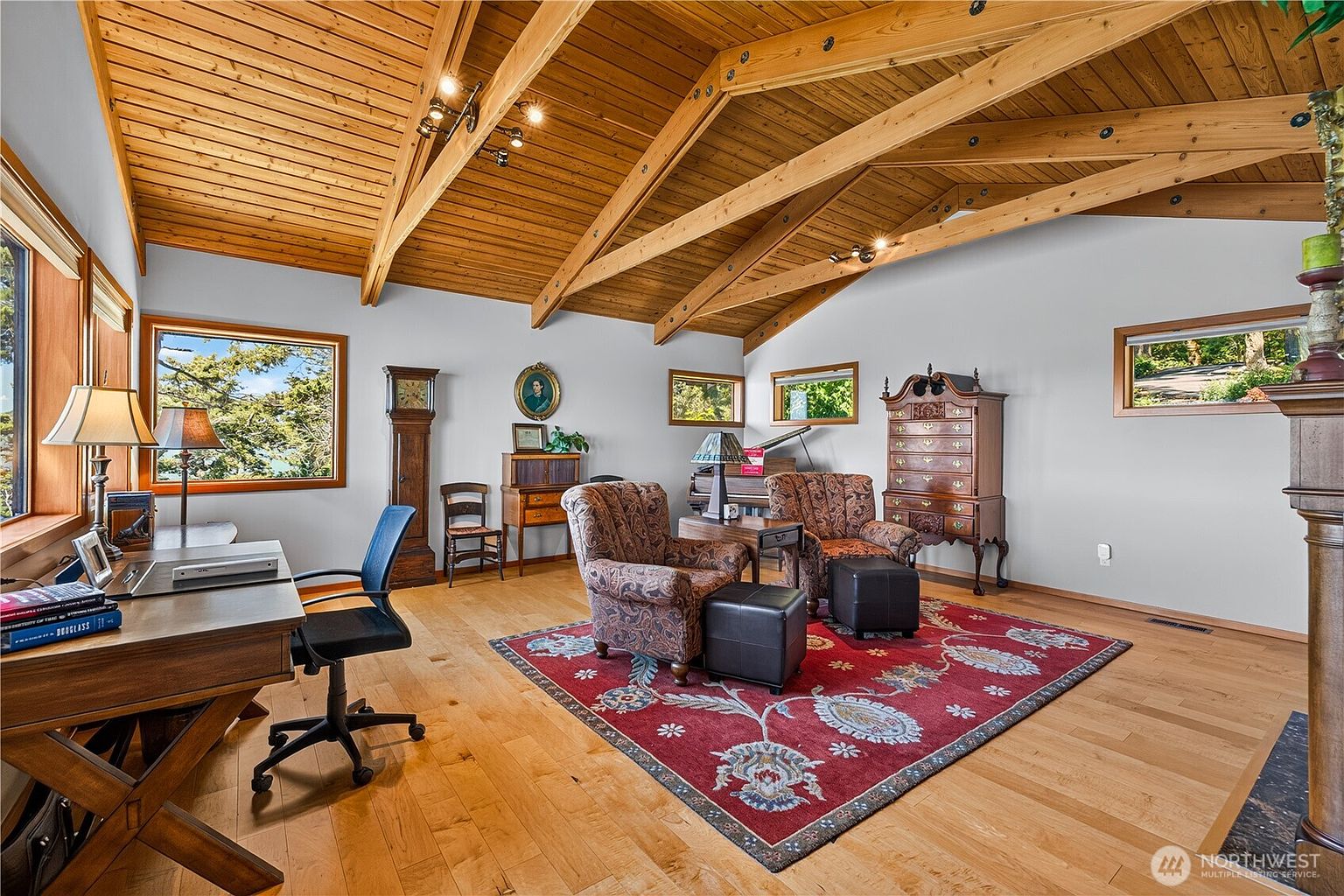 This spacious home office features a vaulted wood-paneled ceiling with exposed beams and light wood flooring. The room is furnished with a large wooden desk, two patterned armchairs with matching ottomans, and a tall antique-style clock, all centered around a vibrant red area rug. Large windows provide ample natural light and views of the surrounding greenery, creating a warm and traditional atmosphere.
