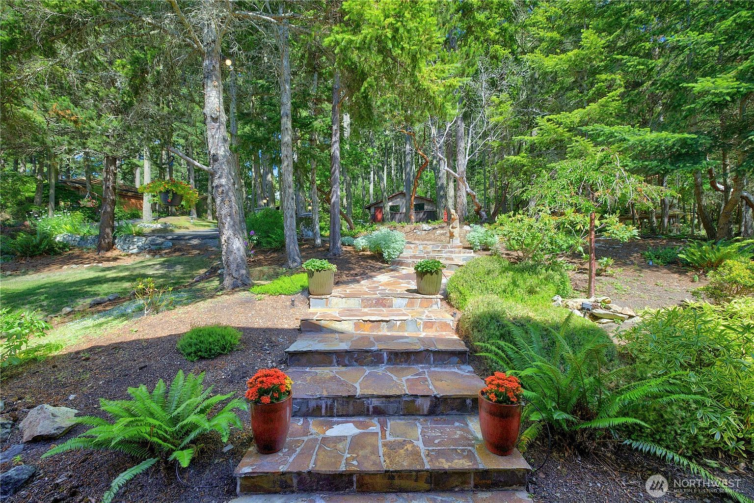 This serene outdoor space features a stone-paved pathway leading up a gentle incline through a lush, wooded landscape. The path is framed by vibrant potted flowers and mature trees, creating a peaceful and inviting garden atmosphere. In the background, a small wooden structure is nestled among the greenery, enhancing the rustic and natural appeal of the property.