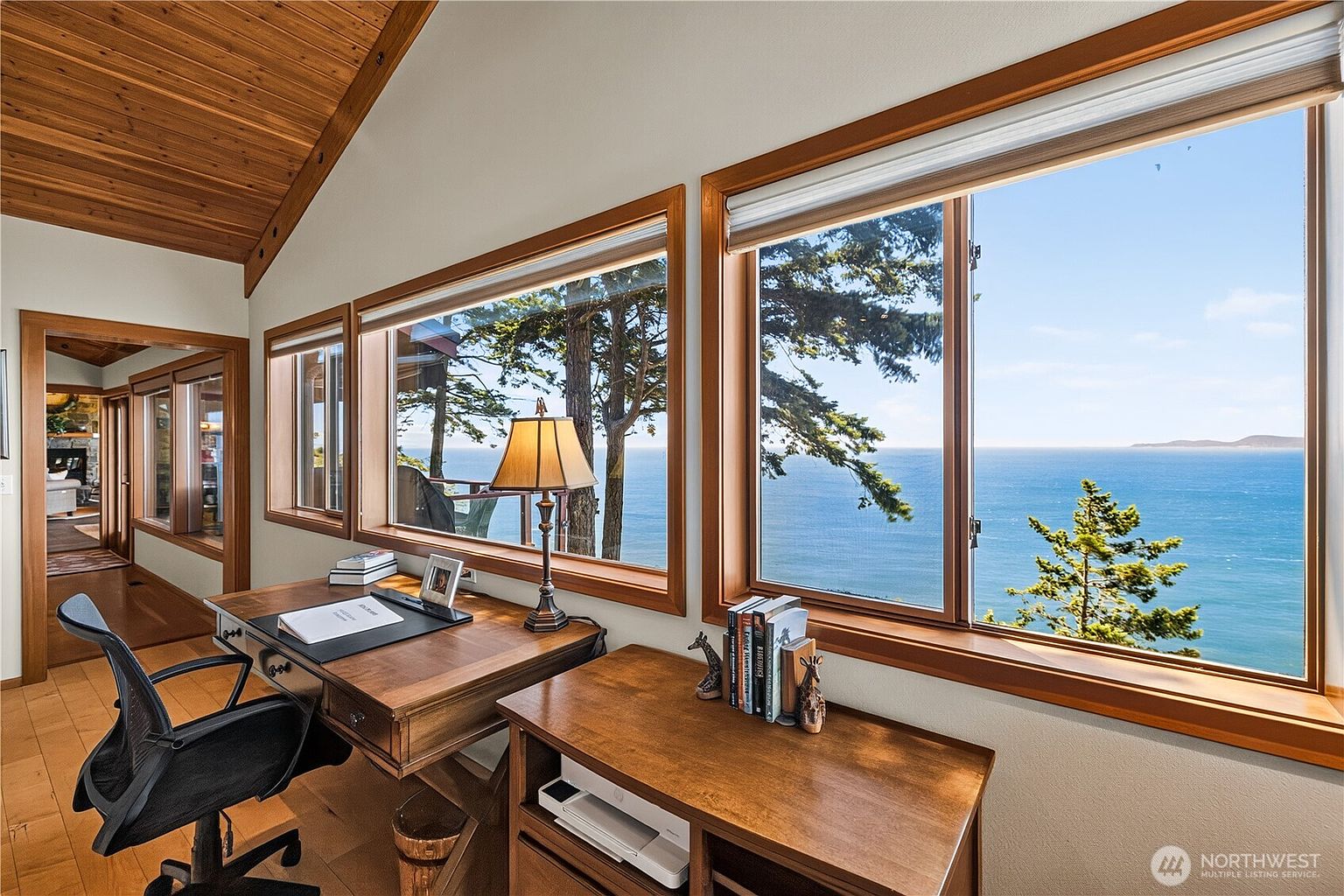 This home office features a warm, wood-toned aesthetic with a vaulted ceiling and large windows that frame a stunning, expansive view of the ocean. A classic wooden desk sits in the foreground, complemented by a black ergonomic office chair and a matching side cabinet holding a printer and books. The room flows into a hallway, providing a glimpse of an adjacent living space, creating an inviting and serene workspace.