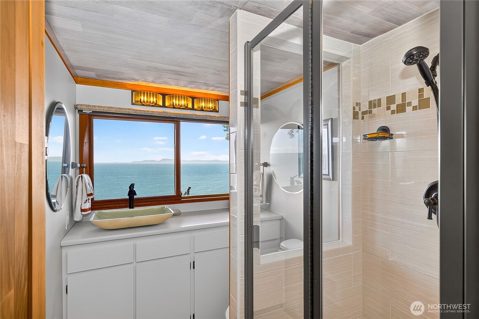 This bathroom features a unique layout with a vanity area positioned directly in front of a large window that offers a panoramic view of the ocean. The space includes a glass-enclosed shower with light-toned wall tiles and a modern showerhead, while a vessel sink sits atop a white vanity cabinet. The perspective captures the seamless transition between the functional bathroom space and the scenic coastal backdrop.