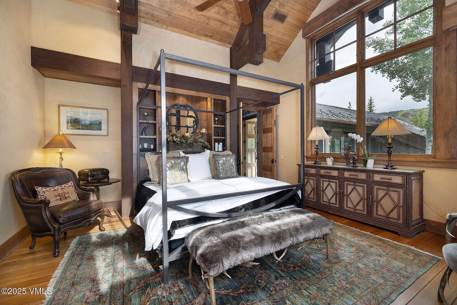 This is a luxurious primary bedroom featuring a four-poster bed with a dark metal frame, complemented by a wooden headboard with built-in shelving and decorative elements. The room is well-lit with natural light from a large window and accented by lamps on a wooden dresser. The overall style is rustic and elegant, creating a warm and inviting atmosphere.