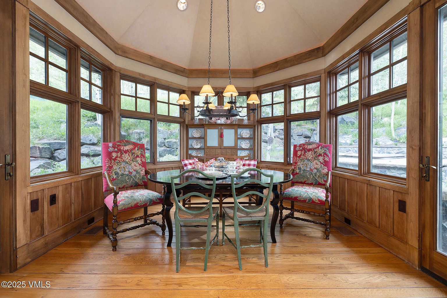 This is an interior shot of a dining room featuring a round table set for a meal, surrounded by chairs with floral upholstery and green painted wood. The room is enclosed by wood-paneled walls and large windows offering natural light and views of the outdoors. A chandelier hangs above the table, and a built-in cabinet displays decorative plates, creating a cozy and elegant dining atmosphere.