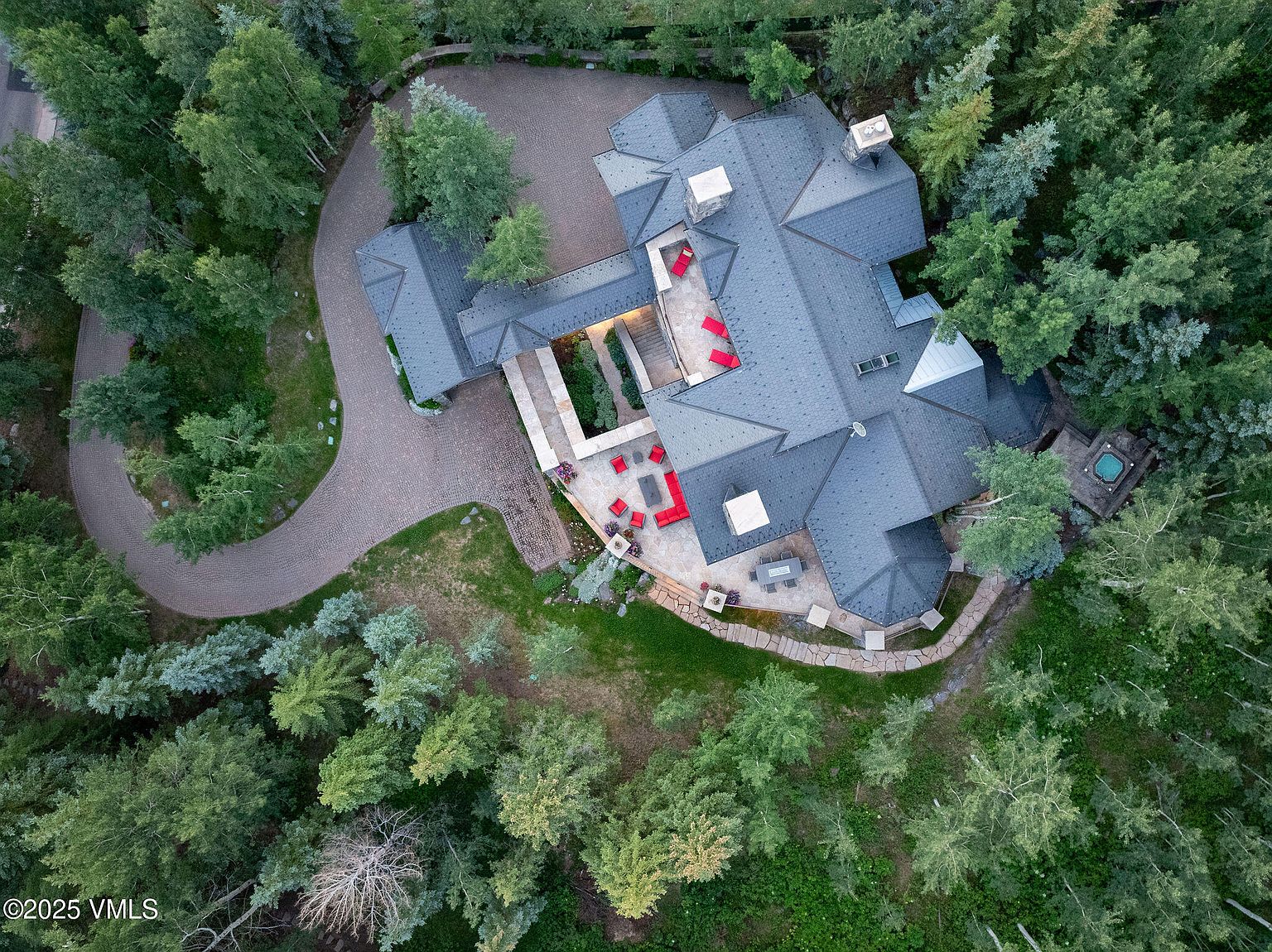 This aerial view showcases a luxurious home nestled among lush trees. The property features a winding driveway, multiple patios with vibrant red furniture, and a unique architectural design with a dark gray roof. A spa is visible on the side of the house, surrounded by trees, adding to the property's secluded and upscale feel.