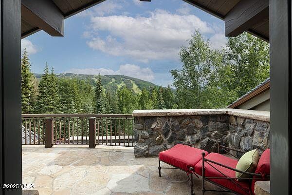 This image showcases a beautiful patio or balcony with stone flooring and a stone wall, offering a serene outdoor living space. A red lounge chair with a green pillow invites relaxation, while the wooden railing provides safety and enhances the rustic charm. The backdrop features lush greenery and a mountain view, creating a tranquil and picturesque setting.