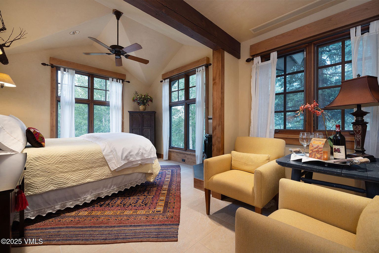 This is a warm and inviting primary bedroom featuring a bed with a yellow patterned quilt and white linens, flanked by windows with sheer white curtains. The room includes two yellow armchairs and a small table with wine and glasses, creating a cozy sitting area. Exposed wooden beams add a rustic touch to the room's design.