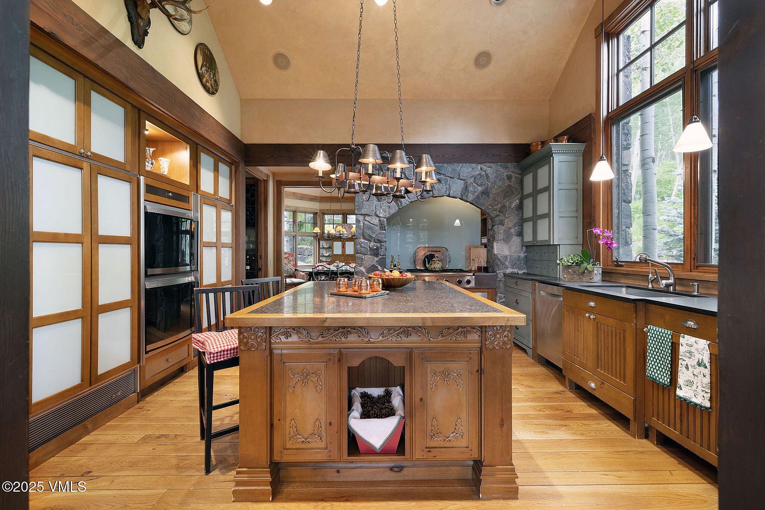 This is a warm and inviting kitchen featuring custom wood cabinetry, a large center island with a dark countertop, and stone accents around the cooking area. The kitchen is well-lit by both natural light from a large window and elegant pendant lighting, creating a cozy and luxurious atmosphere. The hardwood floors add to the rustic charm of the space.