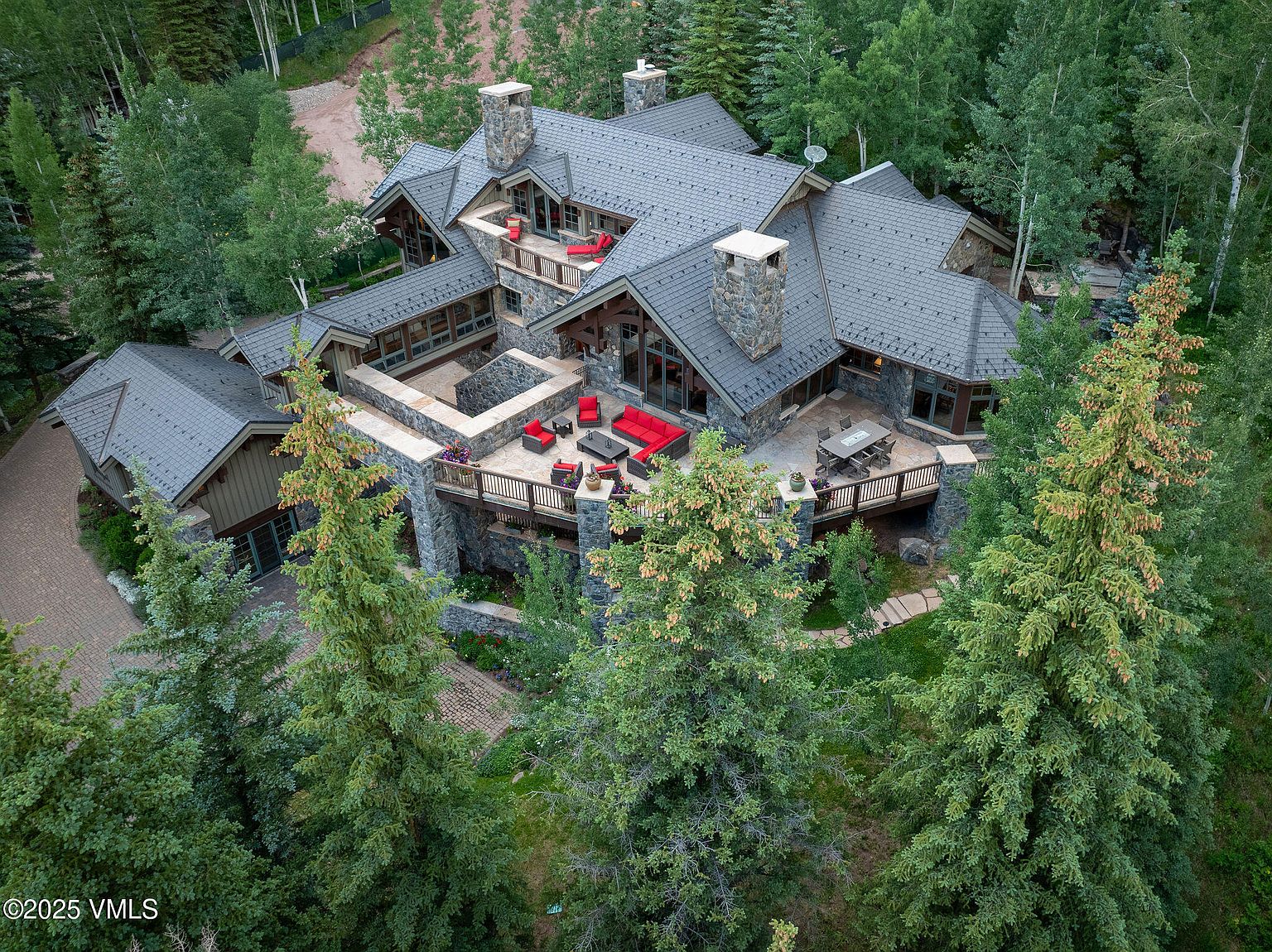 This aerial shot showcases a luxurious, multi-level home nestled among lush trees. The house features a complex roofline with multiple gables, stone chimneys, and expansive patios furnished with outdoor seating. The surrounding landscape provides privacy and a sense of seclusion, making it an ideal retreat.